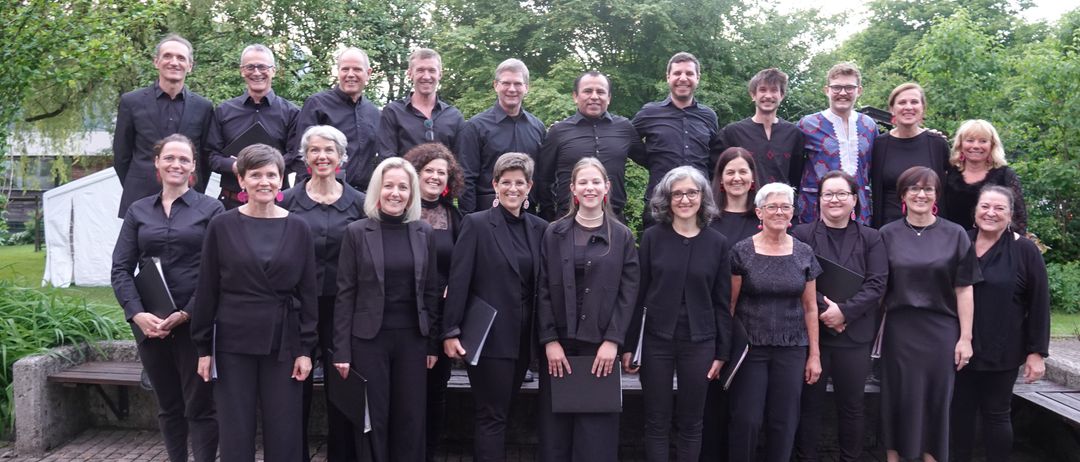 Ein Chor aus Erwachsenen, sowohl Männer als auch Frauen, in Schwarz gekleidet, steht in einem Außenbereich und posiert für ein Foto mit Bäumen im Hintergrund.