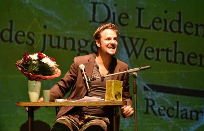 Ein Mann auf der Bühne, lächelnd, mit einem Mikrofon, einem Buch und einer Vase mit Blumen auf einem Tisch vor ihm.