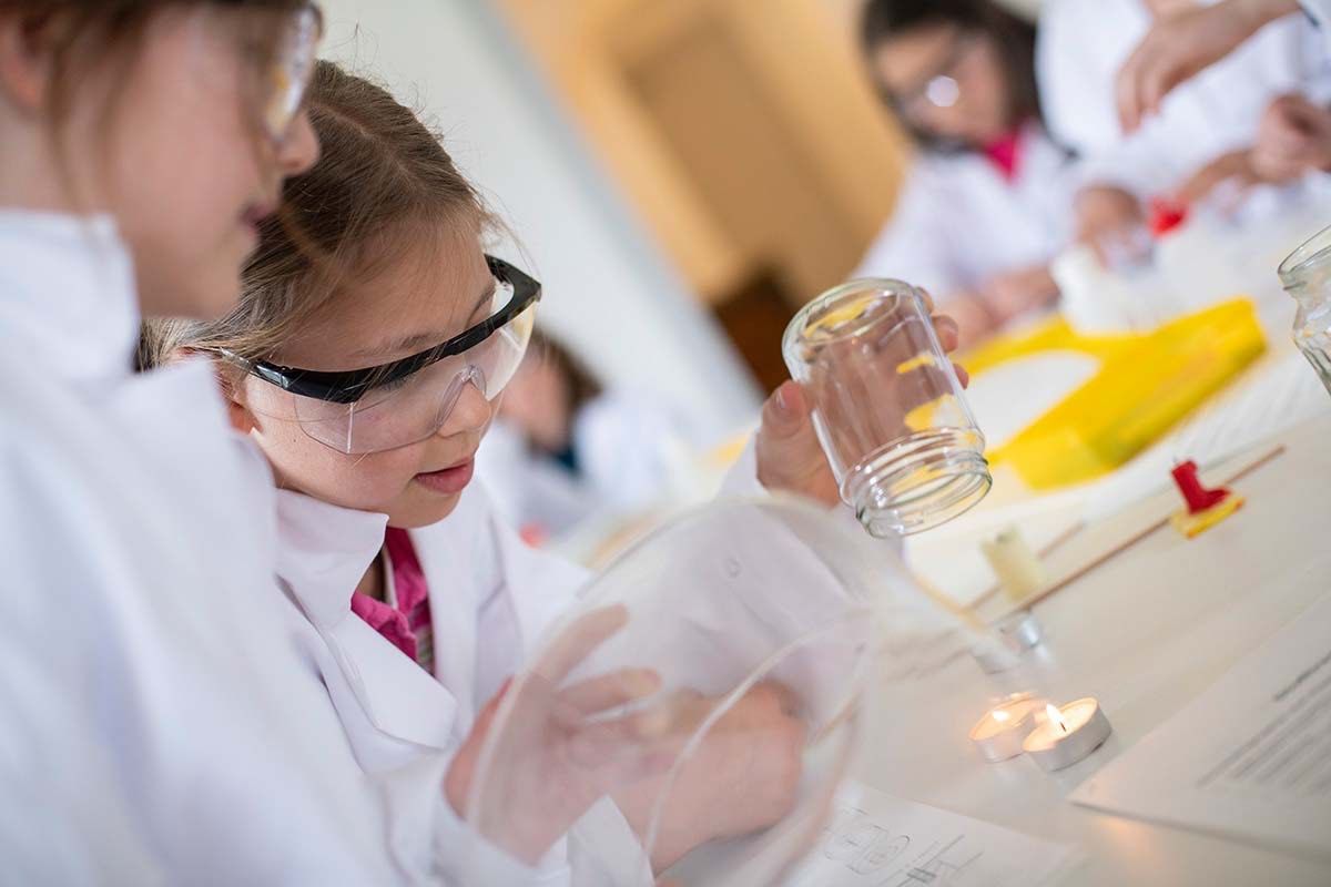 Ein junges Mädchen in einer Laborkittel und Schutzbrille führt ein Experiment mit einem klaren Glasbehälter durch, umgeben von anderen Schülern.