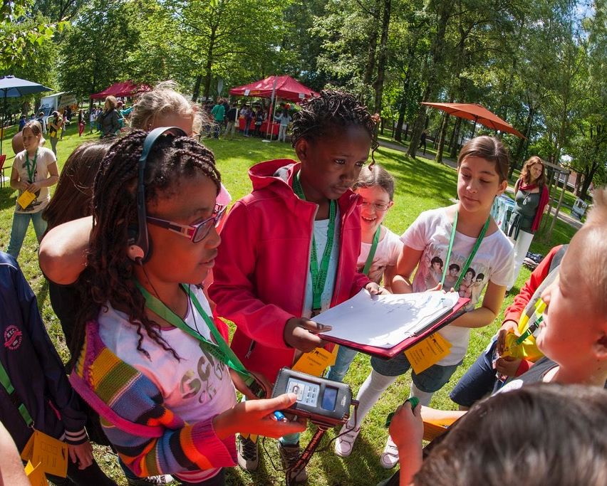 Eine Gruppe von Kindern ist draußen in einem Park und hält ein Buch und ein Gerät. Sie tragen Namensschilder und einige haben Kopfhörer. Ein Mädchen in einer pinken Jacke hält ein Klemmbrett. Bäume und Zelte sind im Hintergrund.