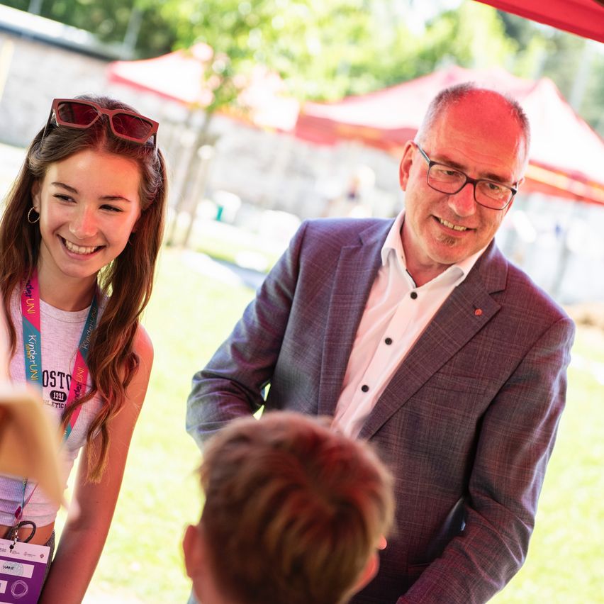 Ein Mann in einem Anzug lächelt und schaut eine junge Frau mit einem Abzeichen an. Das Mädchen lächelt und hat eine Sonnenbrille auf dem Kopf. Sie stehen in einem Außenbereich.