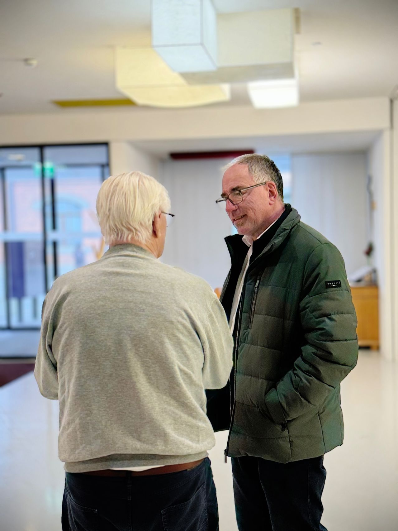 Zwei Männer stehen und unterhalten sich in einem Flur mit Fenstern und einer weißen Decke.