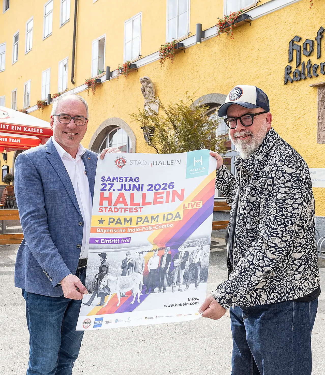 Zwei Männer halten ein Plakat, das für ein Festival in Hallein, Deutschland, wirbt. Das Plakat zeigt eine bayerische Band und den Termin 27. Juni 2026.