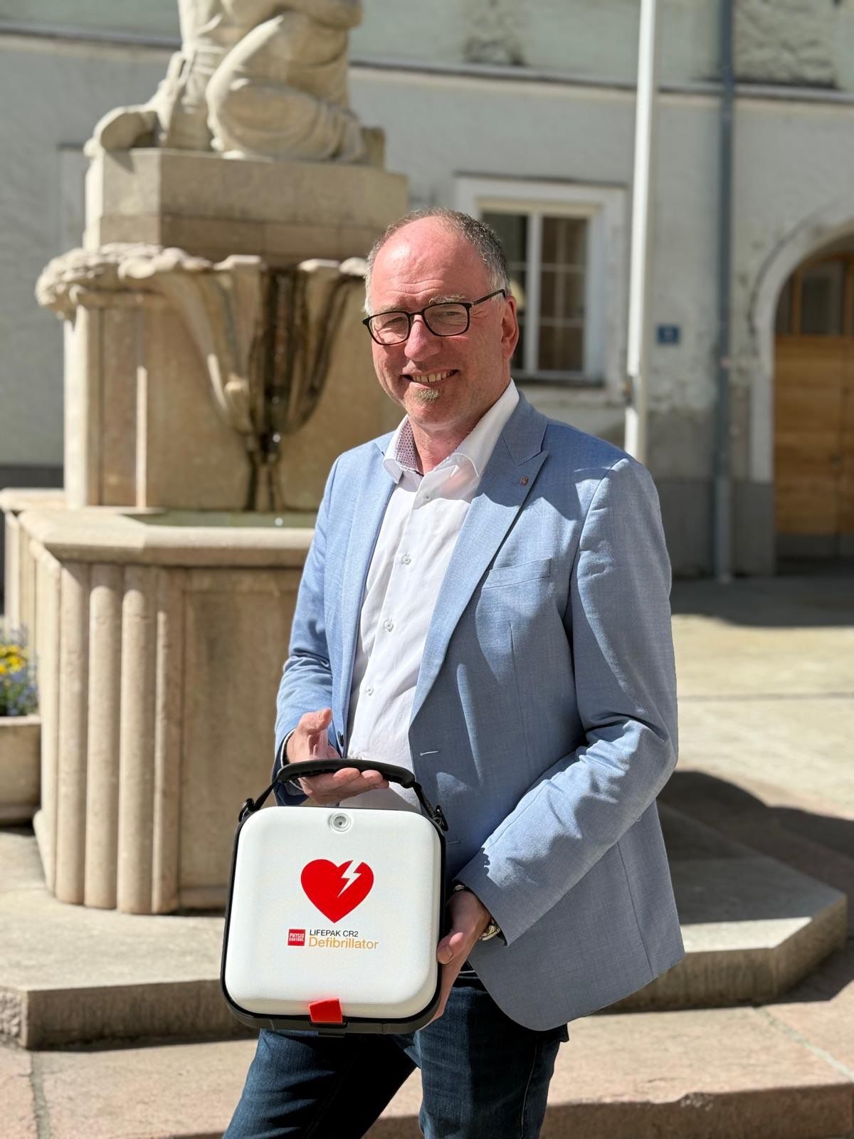 Ein Mann in einem hellblauen Anzug und mit Brille steht im Freien, lächelt und hält ein weiß-rotes Gerät mit Herzsymbol in der Hand.