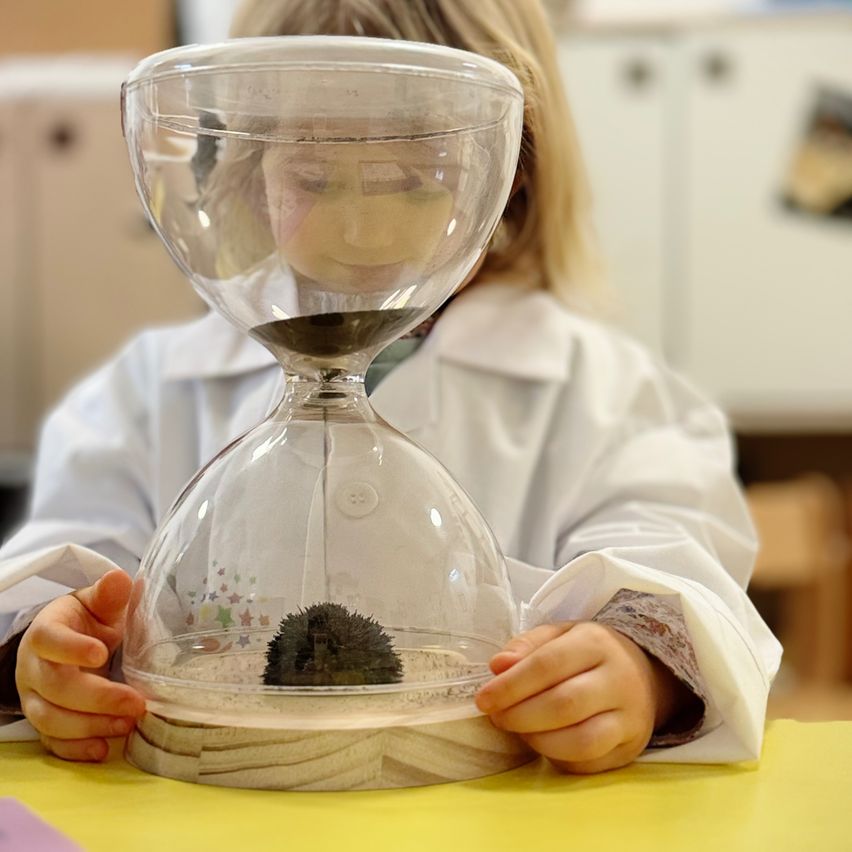 Ein junges Kind in einer Laborkittel hält eine durchsichtige Sanduhr mit einem Igel darin.