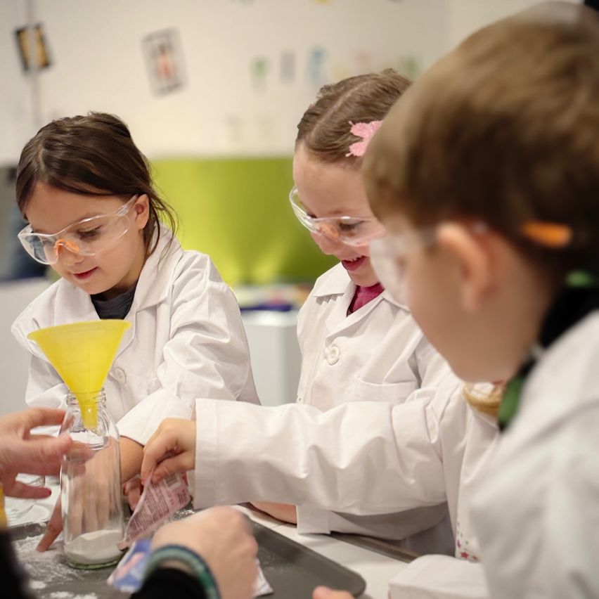 Drei Kinder in Laborkitteln und Schutzbrillen führen ein wissenschaftliches Experiment durch. Eines hält eine gelbe Trichter über einem Glas, ein anderes hält eine Tüte und das dritte beobachtet.
