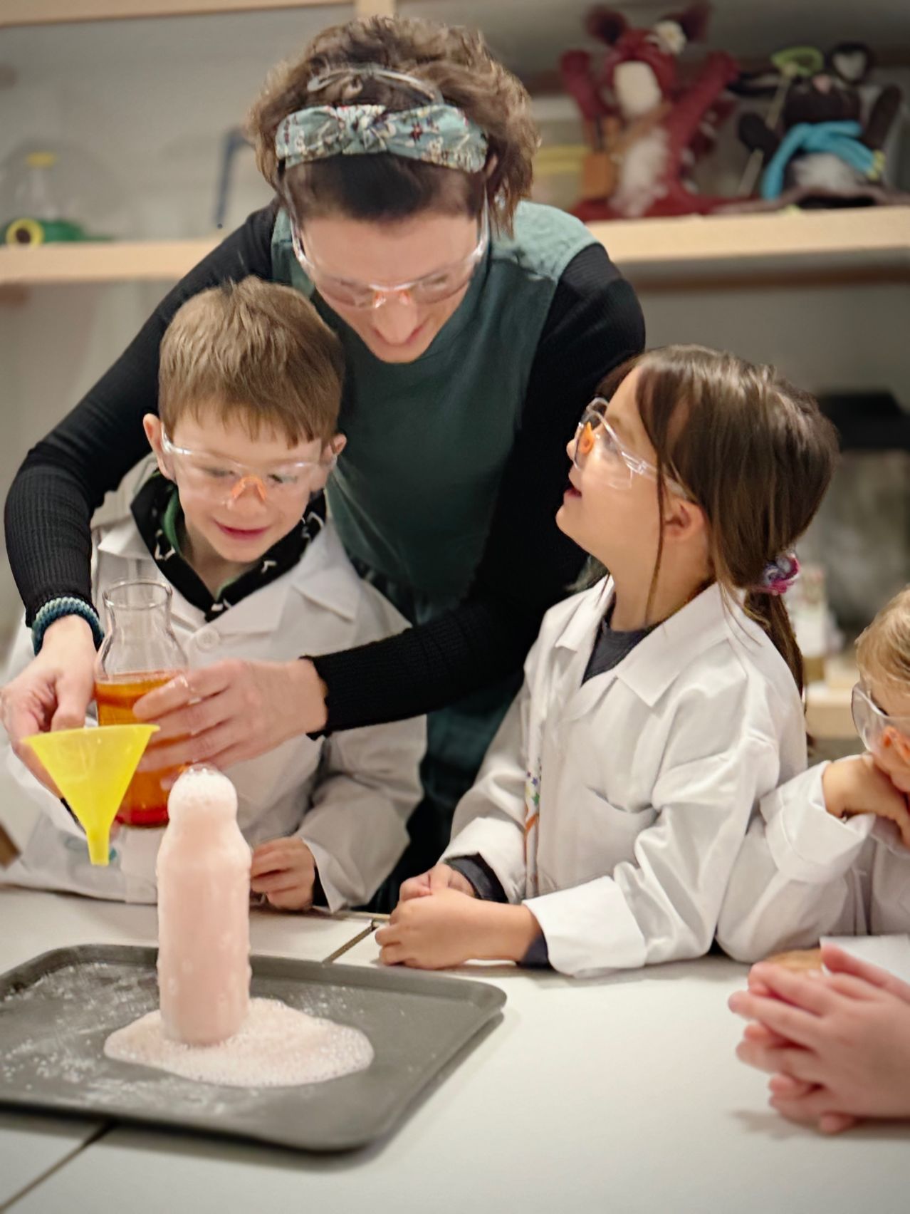 Eine Frau in einer Laborkittel hilft einer Gruppe von Kindern bei einem wissenschaftlichen Experiment. Die Kinder tragen Laborkittel und Schutzbrillen. Eine rosa Flasche mit einem Trichter wird über ein rosa Objekt auf einem Tablett gegossen.