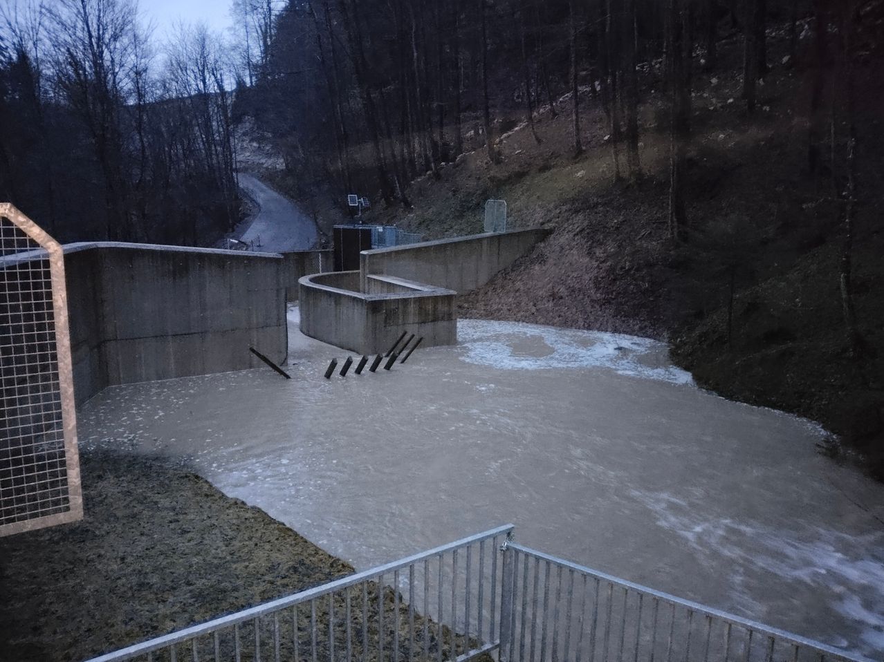 Ein überfluteter Fluss mit einer Betonstaumauerstruktur auf der linken Seite, umgeben von einem Wald. Wasser fließt durch die Staumauer und den Hang hinunter.