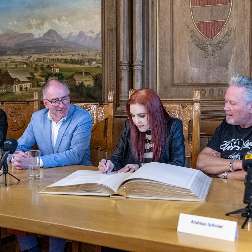 Drei Personen sitzen um einen Tisch herum, wobei eine in ein offenes Buch schreibt. Zwei Männer und eine Frau. Ein Mikrofon steht vor dem Tisch.
