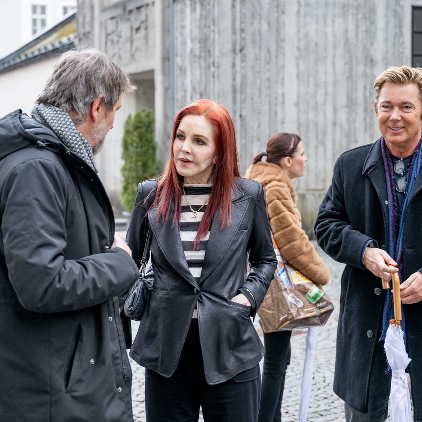 Drei Personen unterhalten sich in der Nähe eines Gebäudes. Eine Frau mit roten Haaren und einer schwarzen Lederjacke steht zwischen zwei Männern, einer hält einen Regenschirm. Dahinter trägt eine Frau in einer Pelzjacke eine große Handtasche.
