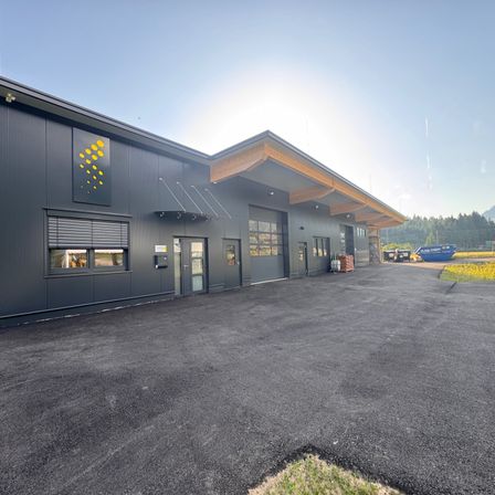 Ein modernes Industriegebäude mit grauer Außenfassade und einem gelben kreisförmigen Design an der Wand. Das Gebäude hat ein geneigtes Dach und große Fenster. Es befindet sich in einem weiten, offenen Bereich mit einem Parkplatz.