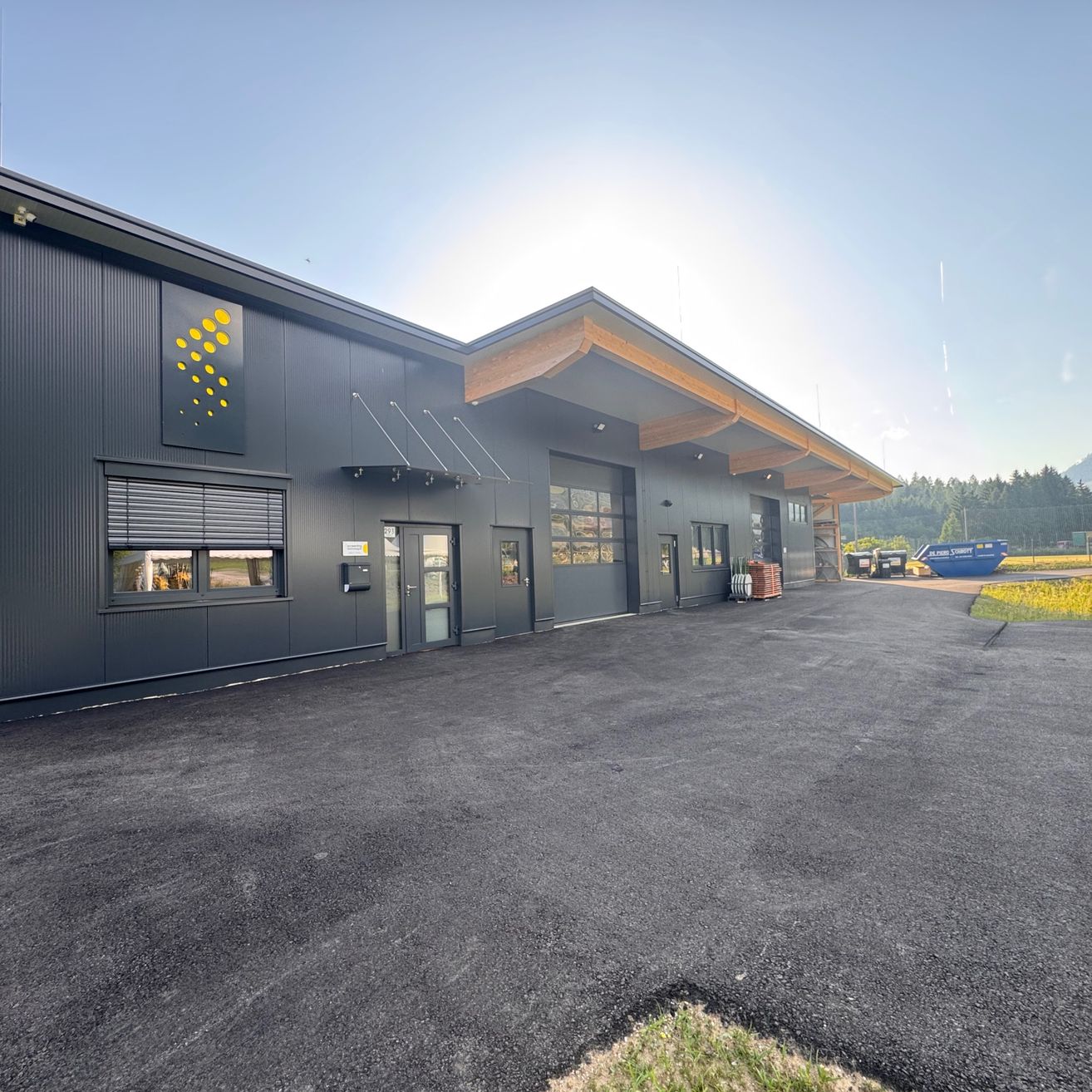 Ein modernes Industriegebäude mit grauer Außenfassade und einem gelben kreisförmigen Design an der Wand. Das Gebäude hat ein geneigtes Dach und große Fenster. Es befindet sich in einem weiten, offenen Bereich mit einem Parkplatz.