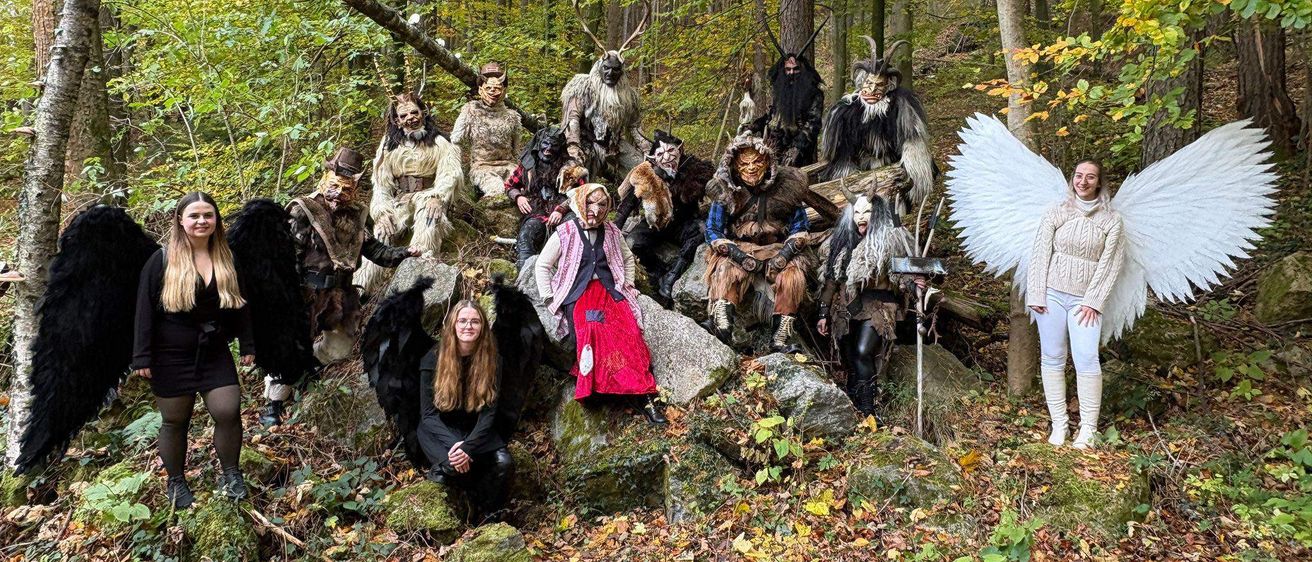 Eine Gruppe von Menschen in Kostümen, möglicherweise für ein Theaterstück oder eine Fotosession, posiert in einem Wald mit gefallenen Blättern und Felsen. Einige tragen Engelsflügel und Masken.