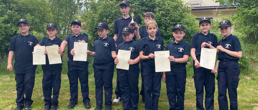 Eine Gruppe von Kindern in gleichen Uniformen steht auf einem Rasen und hält Urkunden in den Händen. Dahinter befindet sich ein Gebäude mit Bäumen und einem Außenbereich. Ein Kind hält einen Pokal.