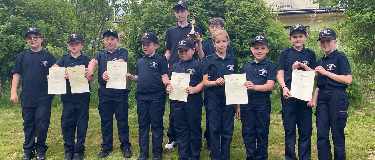 Eine Gruppe von Kindern in gleichen Uniformen steht auf einem Rasen und hält Urkunden in den Händen. Dahinter befindet sich ein Gebäude mit Bäumen und einem Außenbereich. Ein Kind hält einen Pokal.