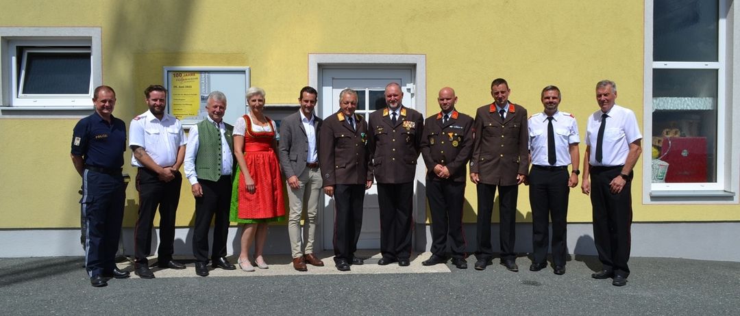 Eine Gruppe von Menschen steht vor einem Gebäude, das Schild sagt Freiwillige Feuerwehr Baumgarten. Sie tragen alle formelle Kleidung.