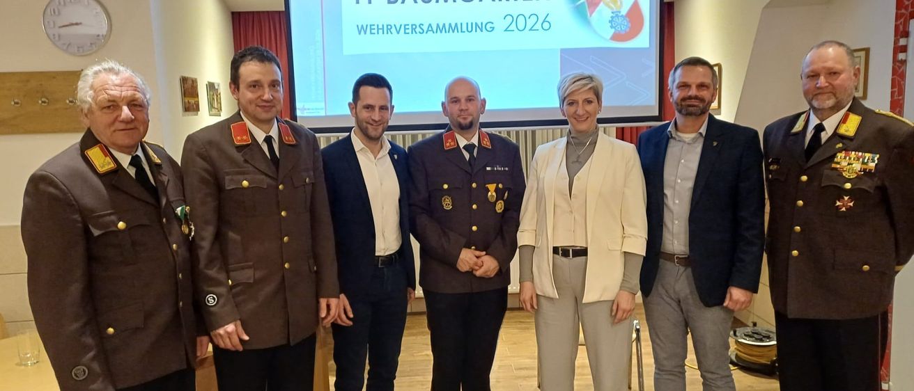 Sieben Personen, darunter drei in Militäruniformen, stehen in einem Konferenzraum vor einer Projektionsleinwand mit dem Text FF BAUMGARTEN WEHRVERSAMMLUNG 2026.