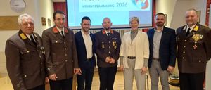 Sieben Personen, darunter drei in Militäruniformen, stehen in einem Konferenzraum vor einer Projektionsleinwand mit dem Text FF BAUMGARTEN WEHRVERSAMMLUNG 2026.