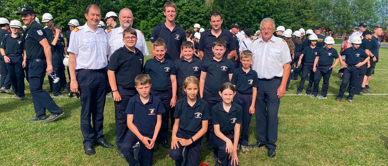 Eine Gruppe junger Menschen in Uniformen steht mit Erwachsenen auf einem Rasenfeld mit Helmen und Trophäen. Sie lächeln und posieren für ein Foto.