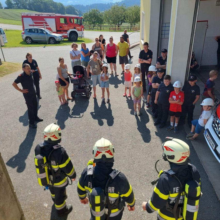 Feuerwehrleute und Kinder stehen um einen Feuerwehrwagen herum. Einige Kinder tragen Helme. Erwachsene und Kinder versammeln sich auf einem Parkplatz. Im Hintergrund sind Fahrzeuge geparkt.