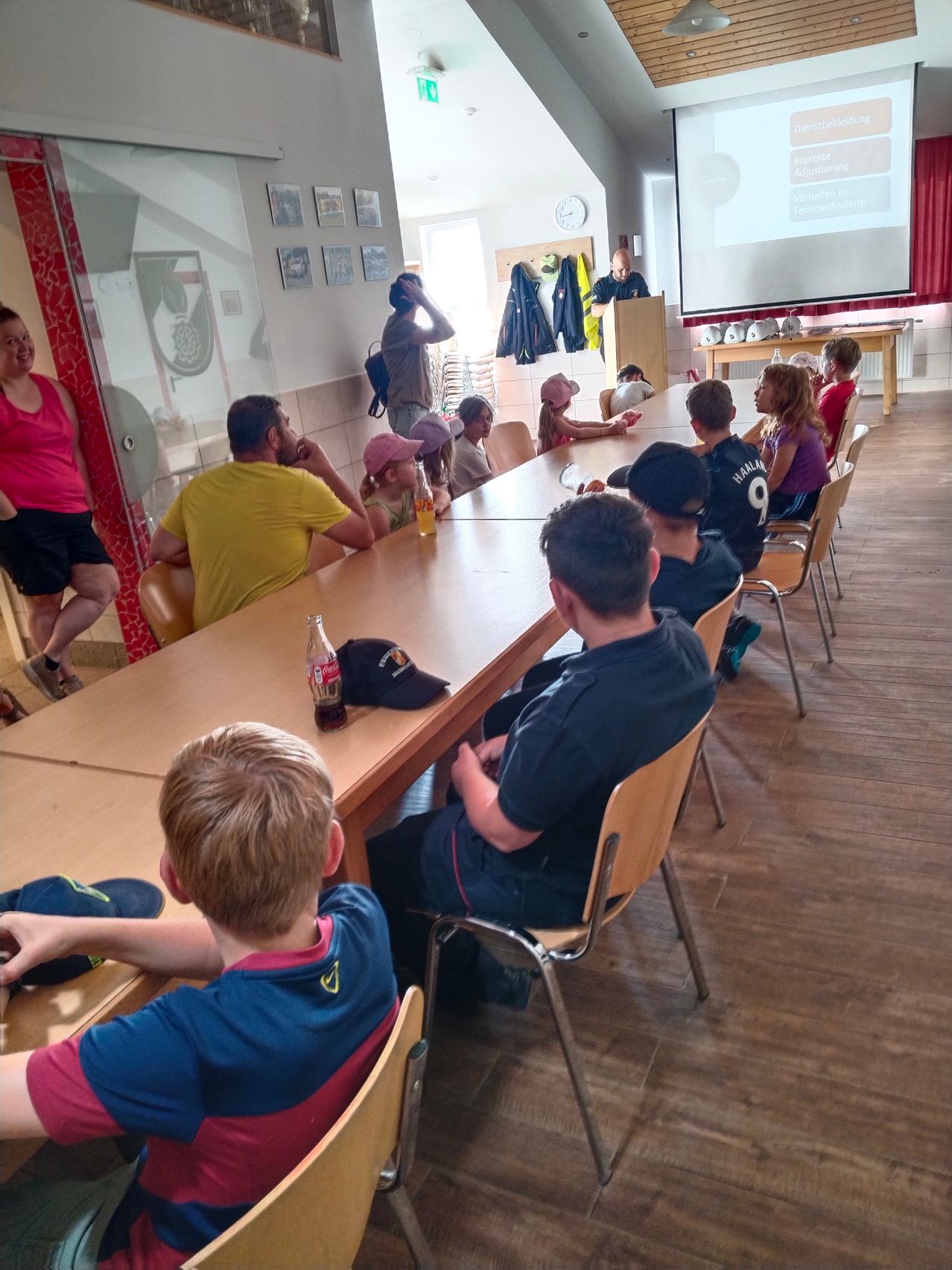 Eine Gruppe von Kindern und Erwachsenen sitzt um einen langen Tisch. Hinter ihnen steht eine Projektionsleinwand und ein Mann steht an einem Podium. Eine Erwachsene steht neben einer Glaswand.