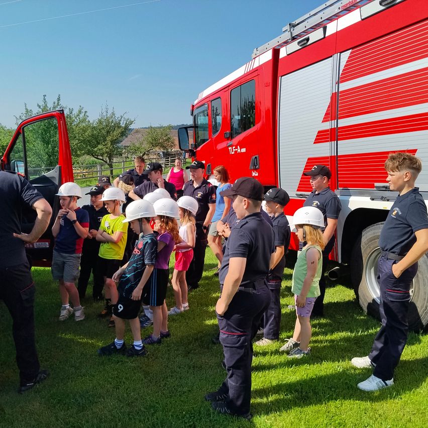 Eine Gruppe von Kindern und Erwachsenen, die Helme tragen, steht vor einem Feuerwehrauto an einem sonnigen Tag. Einige von ihnen machen Fotos.
