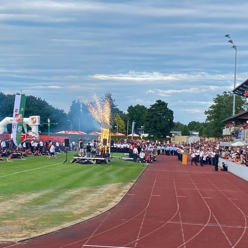 Eine große Menschenmenge versammelt sich auf einem Sportplatz mit einer roten Laufbahn für eine Veranstaltung, mit Feuerwerk, Fahnen und einer Bühne mit einer Band. Bäume und ein bewölkter Himmel bilden einen malerischen Hintergrund.