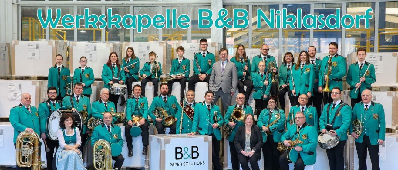 Ein Blasorchester posiert für ein Foto vor einem großen Banner mit der Aufschrift B&B Niklo und einer Box mit der Aufschrift B&B Paper Solutions. Die Mitglieder in grünen Uniformen halten verschiedene Instrumente.