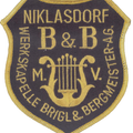 Das Bild zeigt ein schildförmiges Abzeichen mit einer goldenen Leier in der Mitte. Der Text lautet 'NIKLASDORF B&B', darunter steht 'WERKSKAPELLE BRIGL & BERGMEISTERLAG'. Der Rand ist gelb eingefasst.