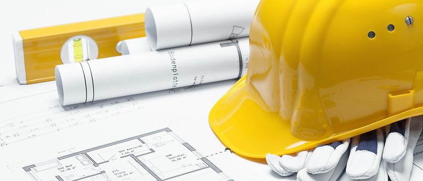 Ein gelber Schutzhelm, Baupläne, ein Taschenrechner, ein Zirkel und Schutzhandschuhe liegen auf einer weißen Oberfläche.