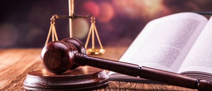 Ein hölzerner Richterhammer liegt auf einem Holztisch neben einem offenen Buch und einer goldenen Waage.