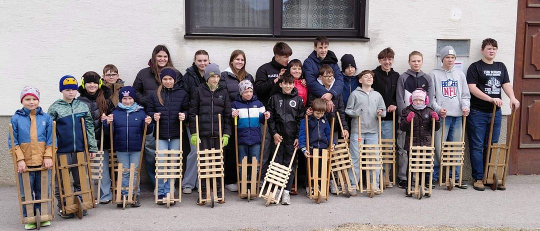 Eine Gruppe von Kindern und Erwachsenen posiert vor einem Gebäude für ein Foto, hält Holzschlitten in den Händen und trägt Winterkleidung.