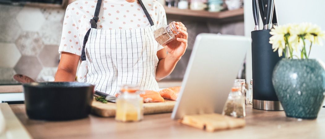 Eine Frau in einer gepunkteten Schürze steht in der Küche und bereitet Essen zu. Sie hält eine Gewürzdose, daneben sind Lachs und ein Messer auf einem Schneidebrett. In der Nähe befinden sich ein Laptop, Schüsseln und Flaschen auf der Arbeitsplatte.