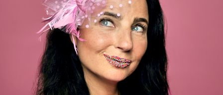 A woman with black hair wearing a pink shirt and a pink feathery headband with a lollipop on it. She has colorful sprinkles on her lips.