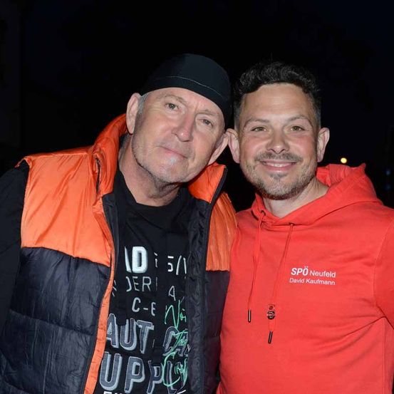Zwei Männer lächeln und posieren für ein Foto. Der Mann auf der linken Seite trägt einen schwarzen Hut und eine orange Weste über einem schwarzen T-Shirt. Der Mann auf der rechten Seite trägt eine rote Kapuzenjacke mit weißem Text, der 'SPD Neufeld David Kaufmann' lautet.