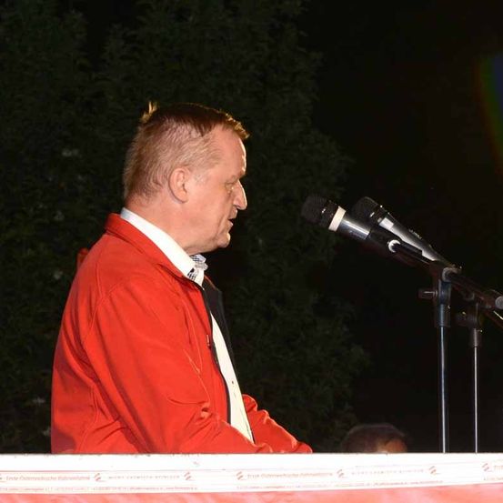 Ein Mann steht auf der Bühne vor einem Mikrofon, gekleidet in eine rote Jacke und ein weißes Hemd.