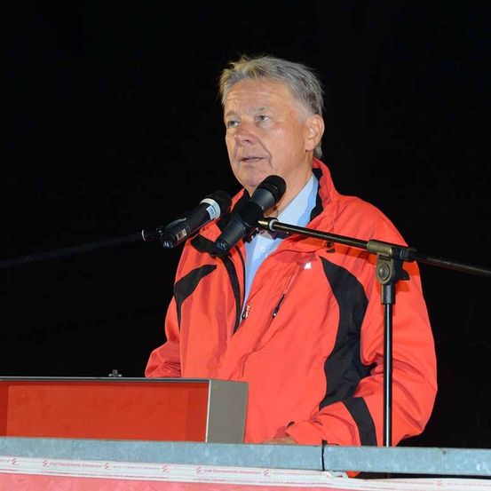 Ein älterer Mann in einer orangefarbenen Jacke steht an einem Rednerpult mit Mikrofon und hält möglicherweise eine Rede.