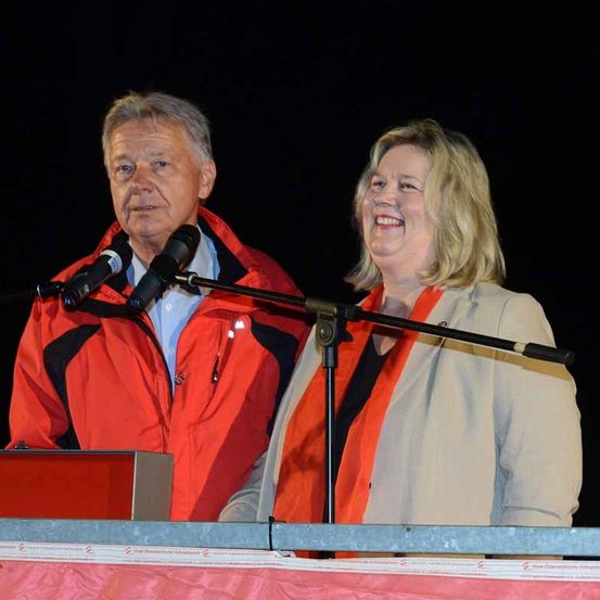 Zwei Personen stehen an einem Rednerpult, einer in einer roten Jacke und der andere in einem beigen Mantel. Sie lächeln beide und schauen etwas vor sich an. Sie befinden sich wahrscheinlich bei einer Veranstaltung.