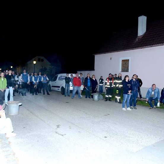 Eine Gruppe von Menschen steht bei Nacht auf der Straße vor einem Haus. Einige Leute tragen Jacken und Mützen, andere Uniformen. Zwei Eimer stehen an der Seite. Ein Van mit dem Wort 'Kilgen' ist vor dem Gebäude geparkt.