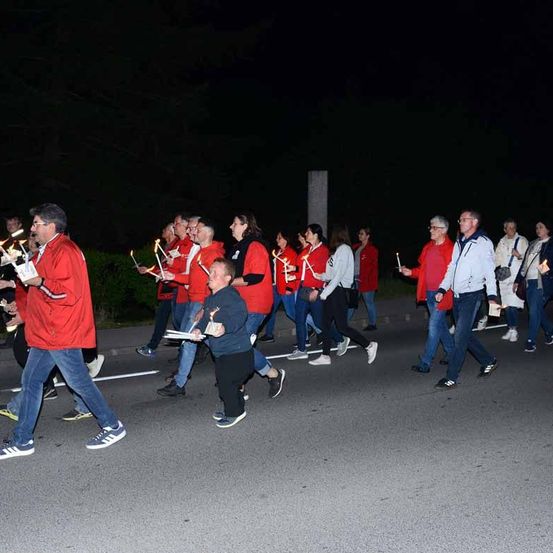 Eine Gruppe von Menschen läuft nachts auf der Straße und hält Kerzen in den Händen. Sie tragen rote Jacken und Turnschuhe.