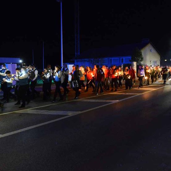 Eine Gruppe von Menschen, die nachts auf einer Straße marschiert, mit brennenden Kerzen in der Hand und Musikinstrumente spielt, beleuchtet durch das Licht der Gebäude und Straßenlaternen.