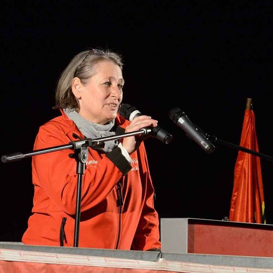 Eine Frau steht auf einer Bühne, hält ein Mikrofon in der Hand und scheint eine Rede zu halten.