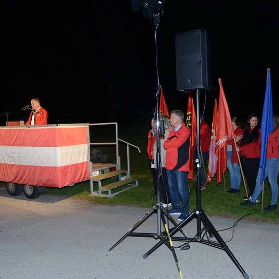 Eine politische Veranstaltung im Freien bei Nacht mit einem Redner am Podium, stehenden Lautsprechern und Teilnehmern in roten Jacken.