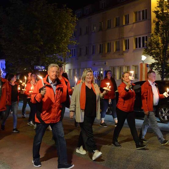 Gruppe von Menschen in roten Jacken und mit Kerzen in der Hand geht bei Nacht die Straße entlang.