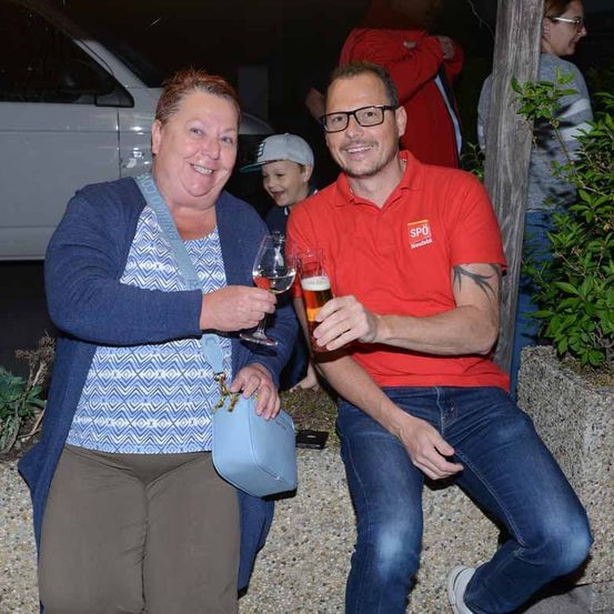 Ein Mann und eine Frau lächeln und halten ein Glas Bier, während sie auf einer Betonbank sitzen.