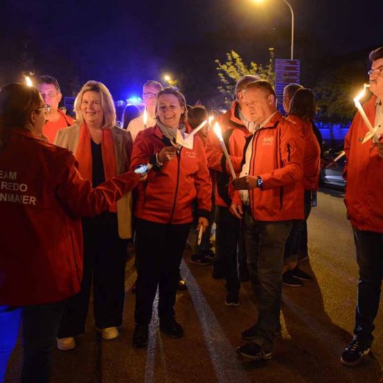 Eine Gruppe von Menschen in roten Jacken mit Kerzen in der Hand geht bei Nacht die Straße entlang.