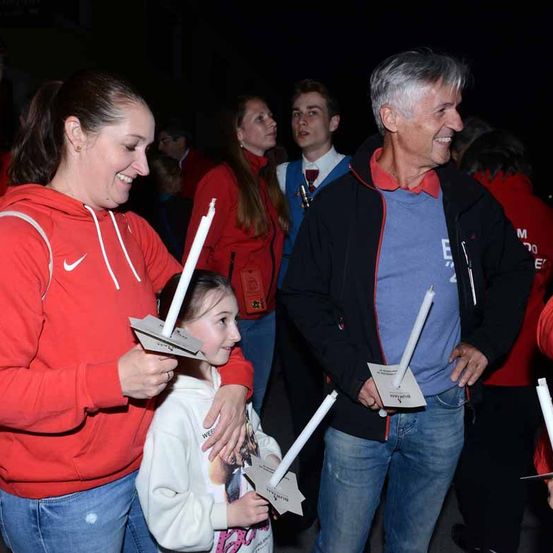 Eine lächelnde Frau, die eine Kerze in einem kerzenbeleuchteten Raum hält, neben ihr ein lächelnder älterer Mann.