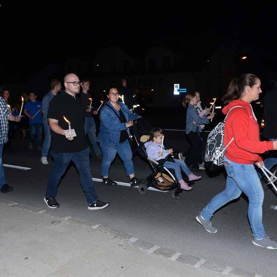 Eine Gruppe von Menschen mit brennenden Kerzen geht bei Nacht auf der Straße. Eine Frau in einem roten Shirt führt die Gruppe an, und ein Kinderwagen mit einem kleinen Kind wird von einer Frau in einer Jeansjacke geschoben.