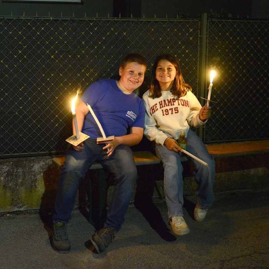 Ein Junge und ein Mädchen sitzen auf einer Bank, lächeln und halten brennende Kerzen. Hinter ihnen ist ein Zaun mit schwarzem Hintergrund zu sehen.