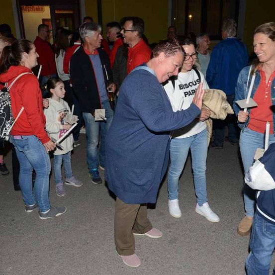Eine Gruppe von Menschen steht im Freien, lächelt und lacht. Eine Frau klatscht und ein Mann hält ein Bier.